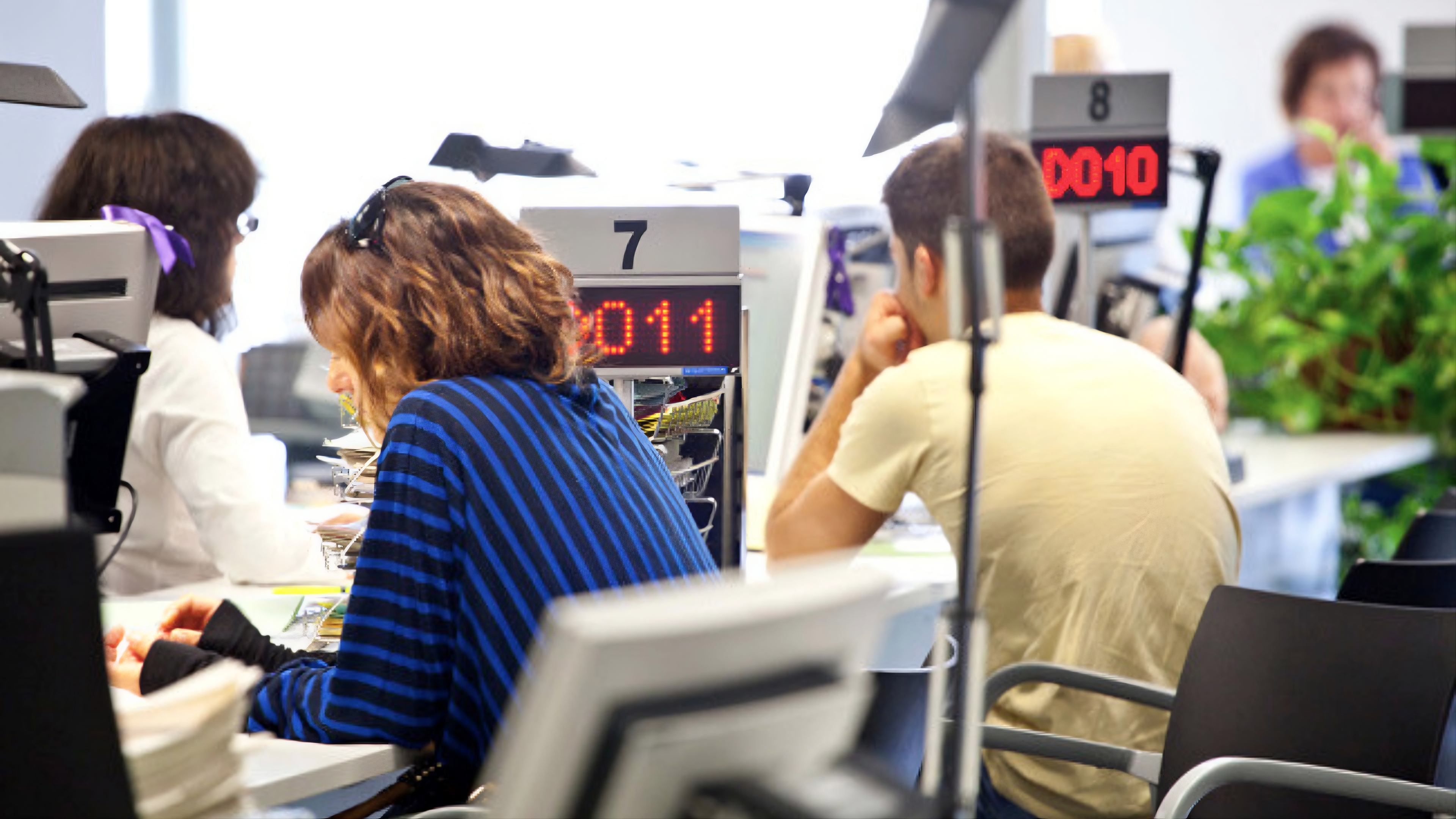 Interior de una Oficina de Empleo con dos personas sentadas frente a puntos de servicio numerados e indicadores de turno digitales en rojo | Gencat