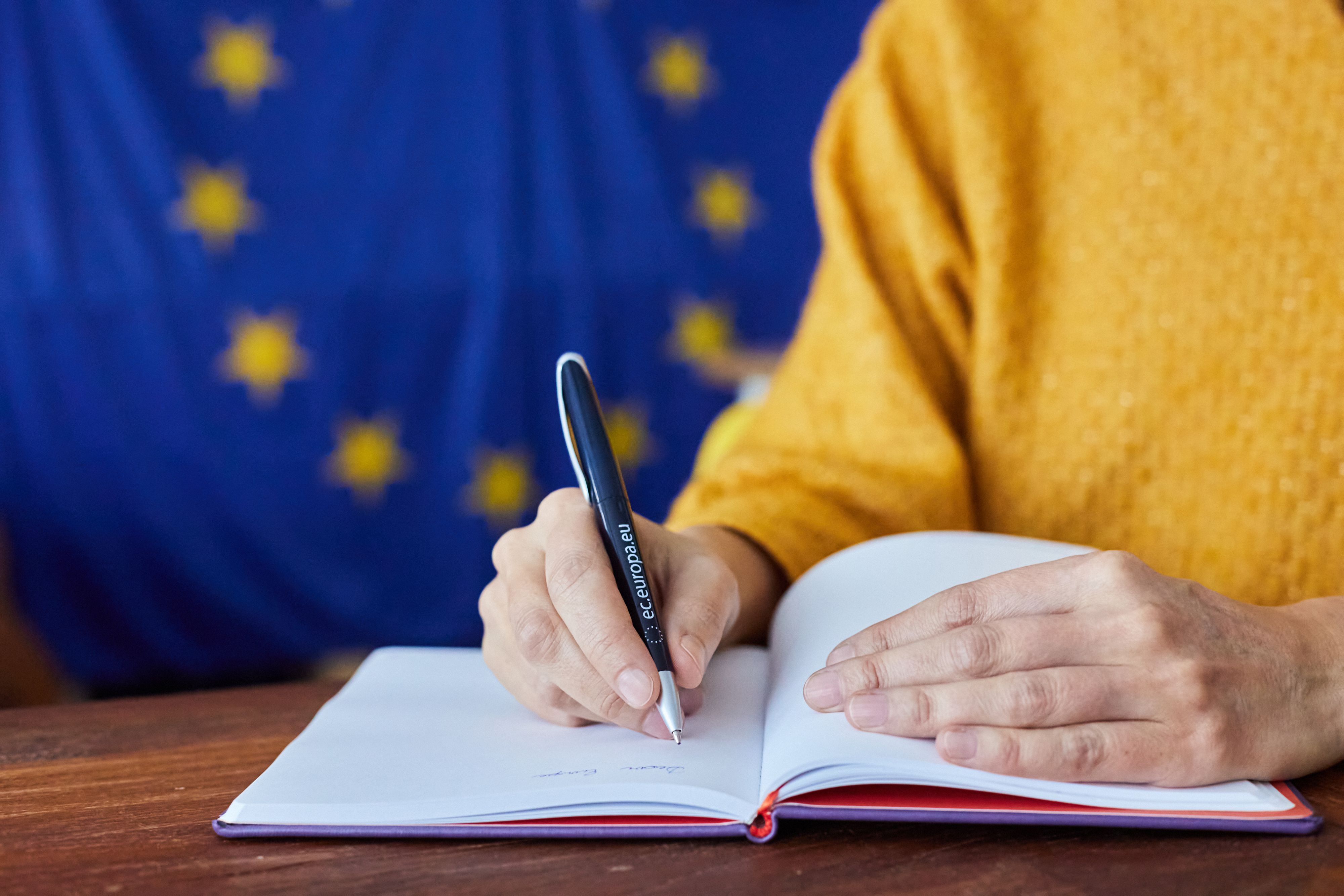 Una persona escrivint en una llibreta amb un bolígraf de la UE i la bandera de la UE al fons | Unió Europea